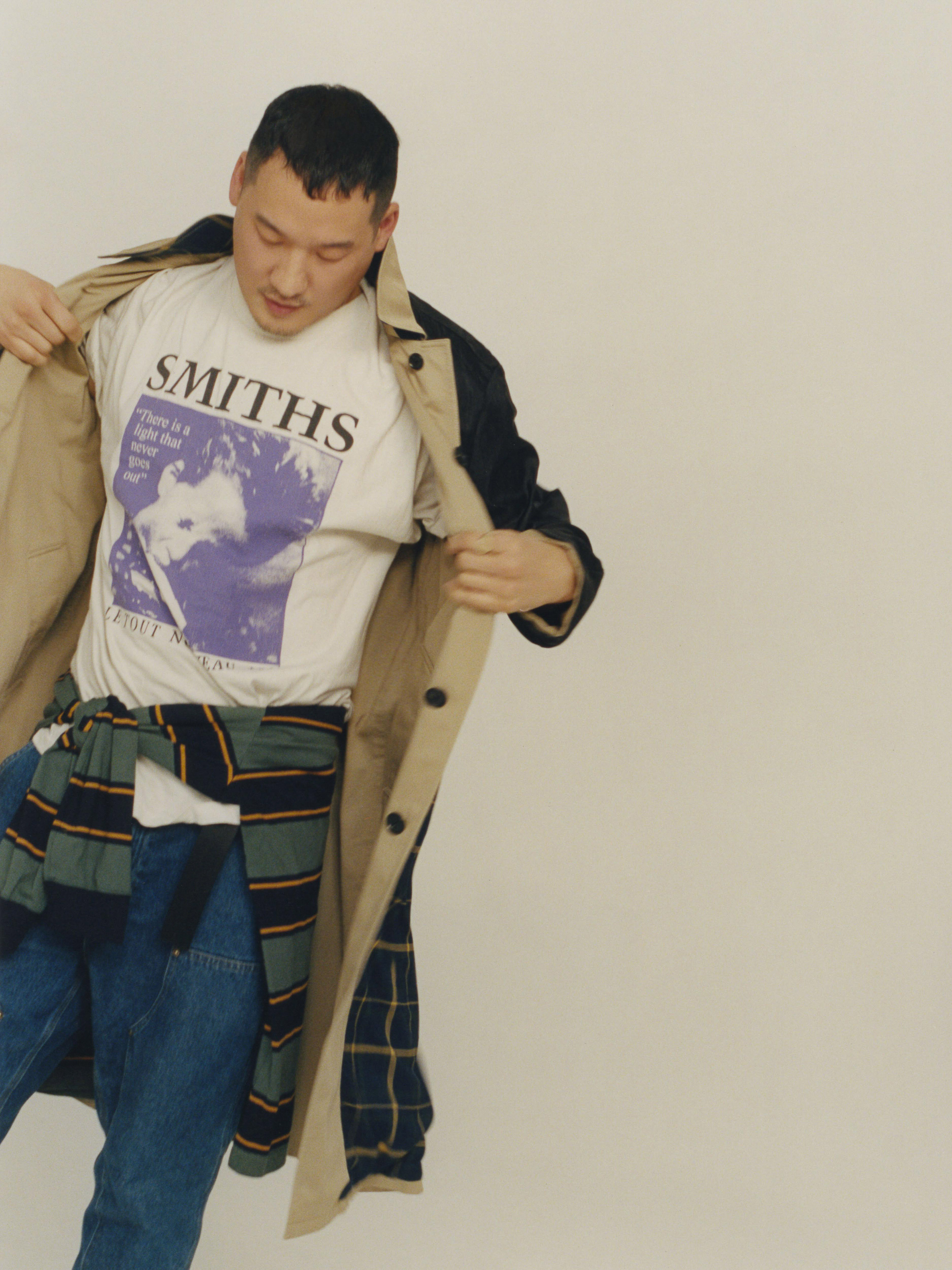 Asian man in an open trench coat with a graphic T-shirt and a striped sweater tied around his waist stands indoors.