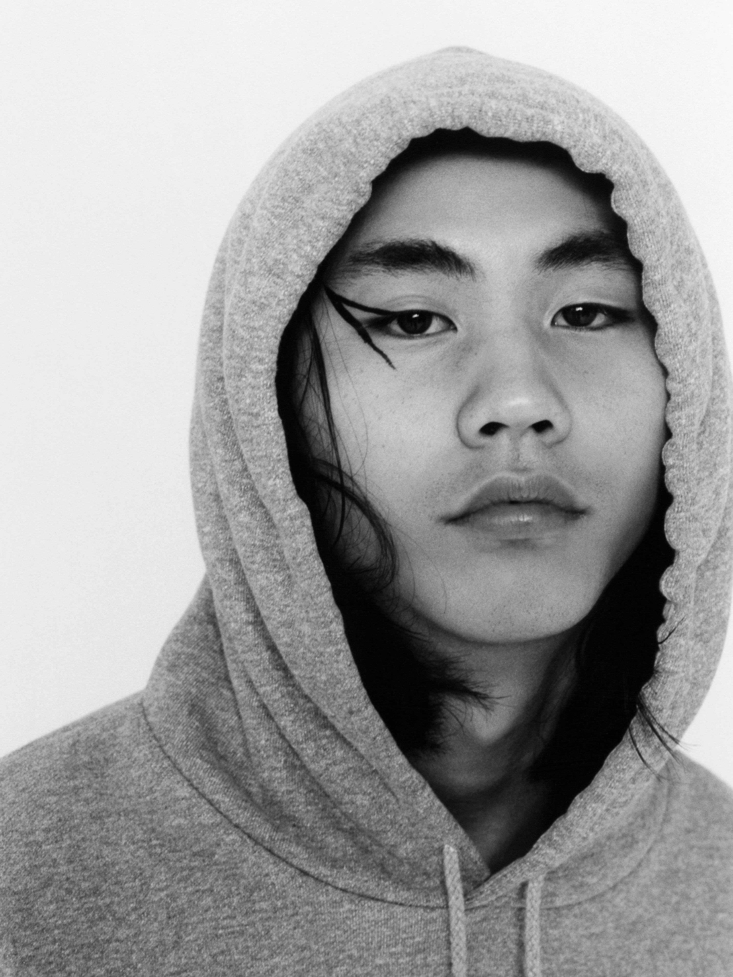 Monochrome portrait of a young Asian adult with winged eyeliner, wearing a gray hoodie against a white backdrop, close-up shot.