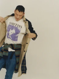 Man wearing a graphic print t-shirt and tan trench coat over blue jeans, captured against a neutral studio backdrop.