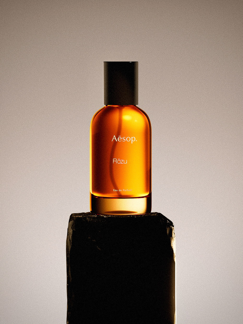 Amber-colored Aesop Rōzu perfume bottle with black cap atop a rough, dark stone against a soft gradient background.