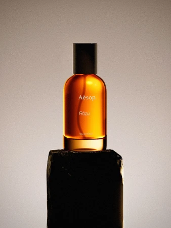Amber-colored Aesop Rōzu perfume bottle with black cap atop a rough, dark stone against a soft gradient background.