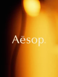 The Aesop brand logo in white serif typography set against an abstract, out-of-focus golden amber and warm glowing background.