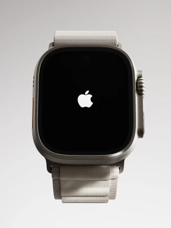 Close-up of an Apple Watch Ultra displaying the Apple logo on a black screen, paired with a neutral-toned fabric band, all against a bright grey backdrop.