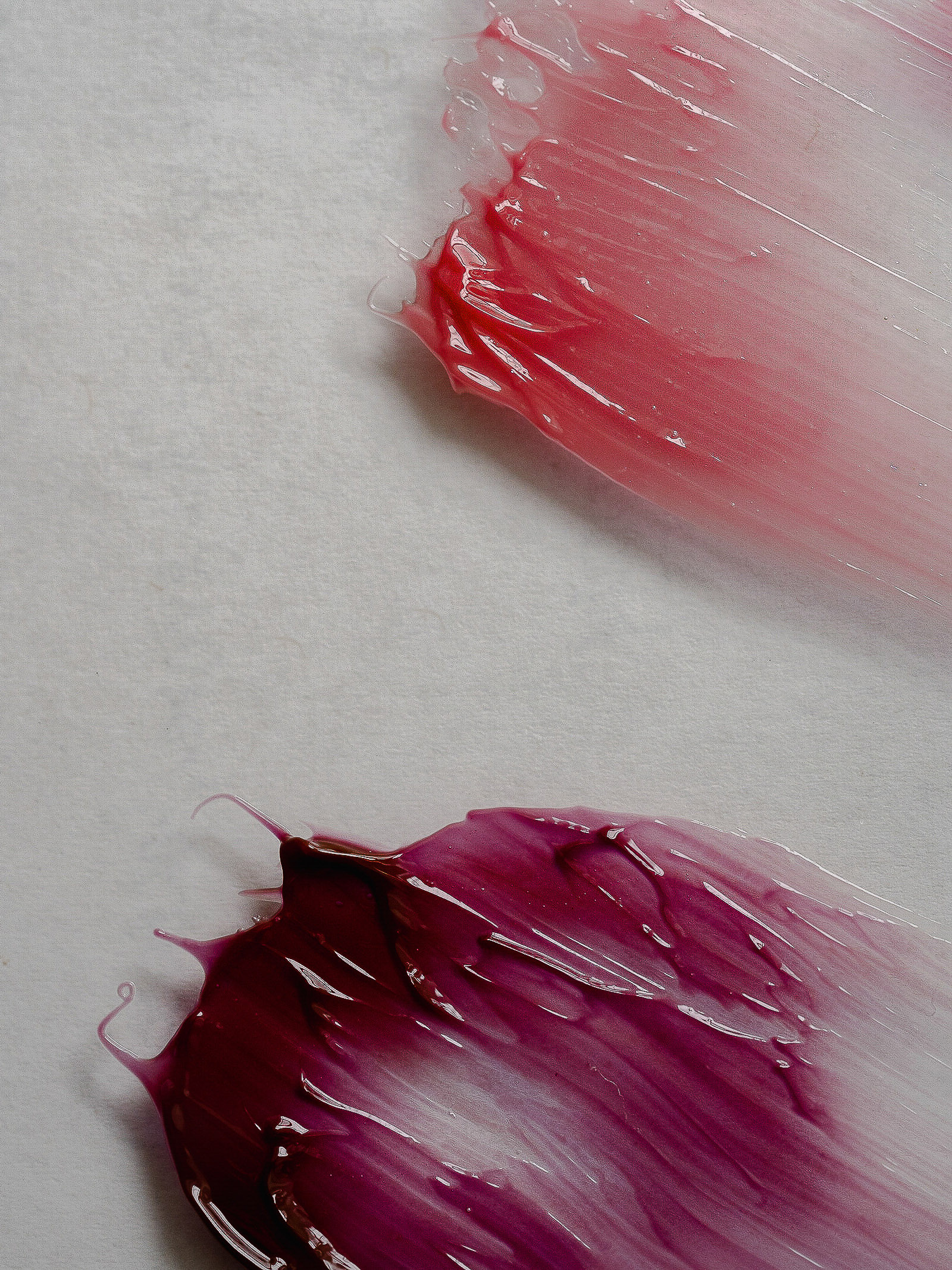 Two dabs of glossy lip gloss on white paper. One dab is reddish pink, and the other is deep magenta and creates a glossy texture.