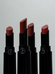 Four open Armani Beauty lipsticks in nude and terracotta shades in black tubes against a light neutral background.
