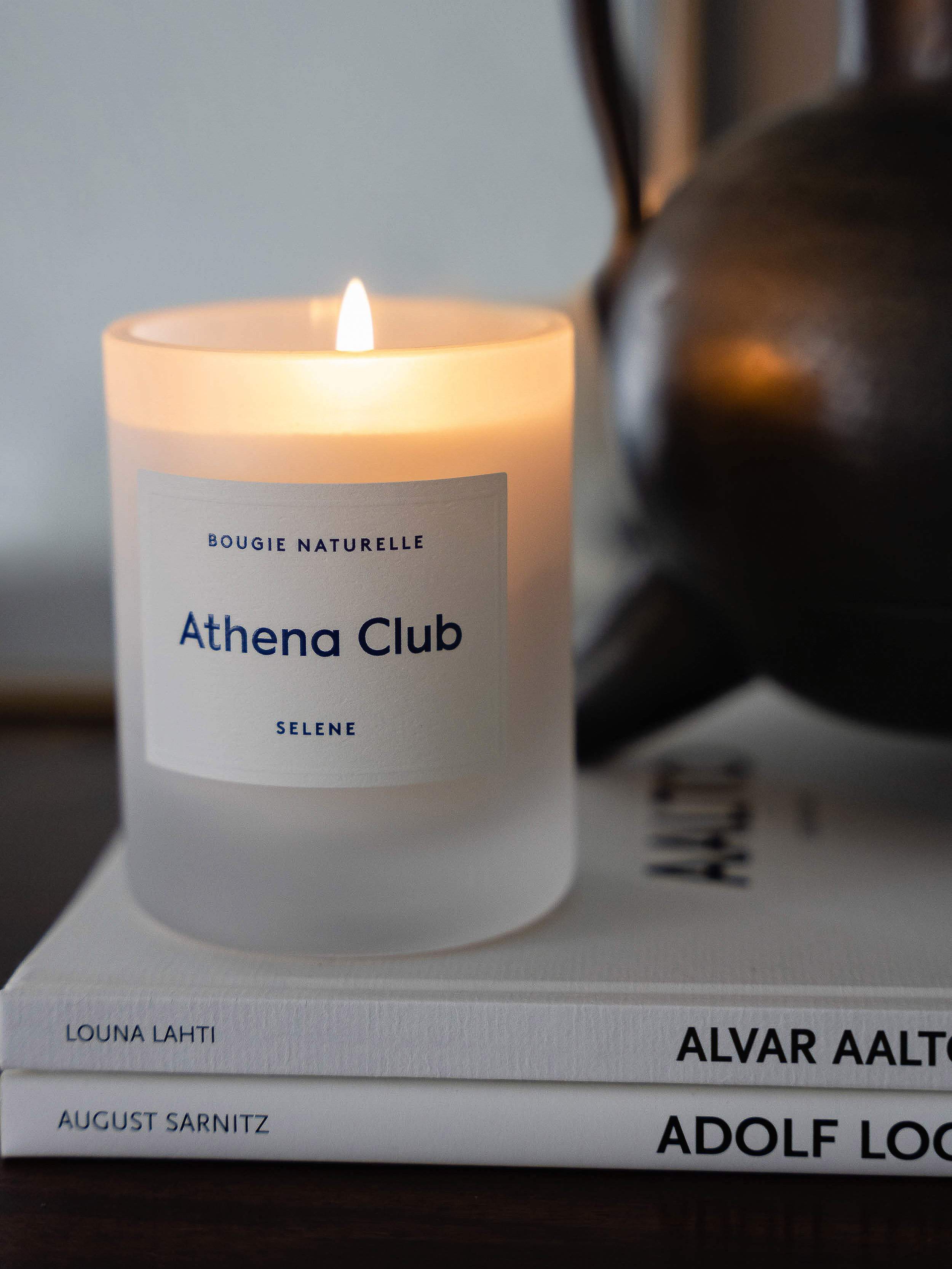 Candle with frosted glass container burning on top of books. The candle label has 'Athena Club' in blue font.