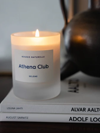 Candle with frosted glass container burning on top of books. The candle label has 'Athena Club' in blue font.