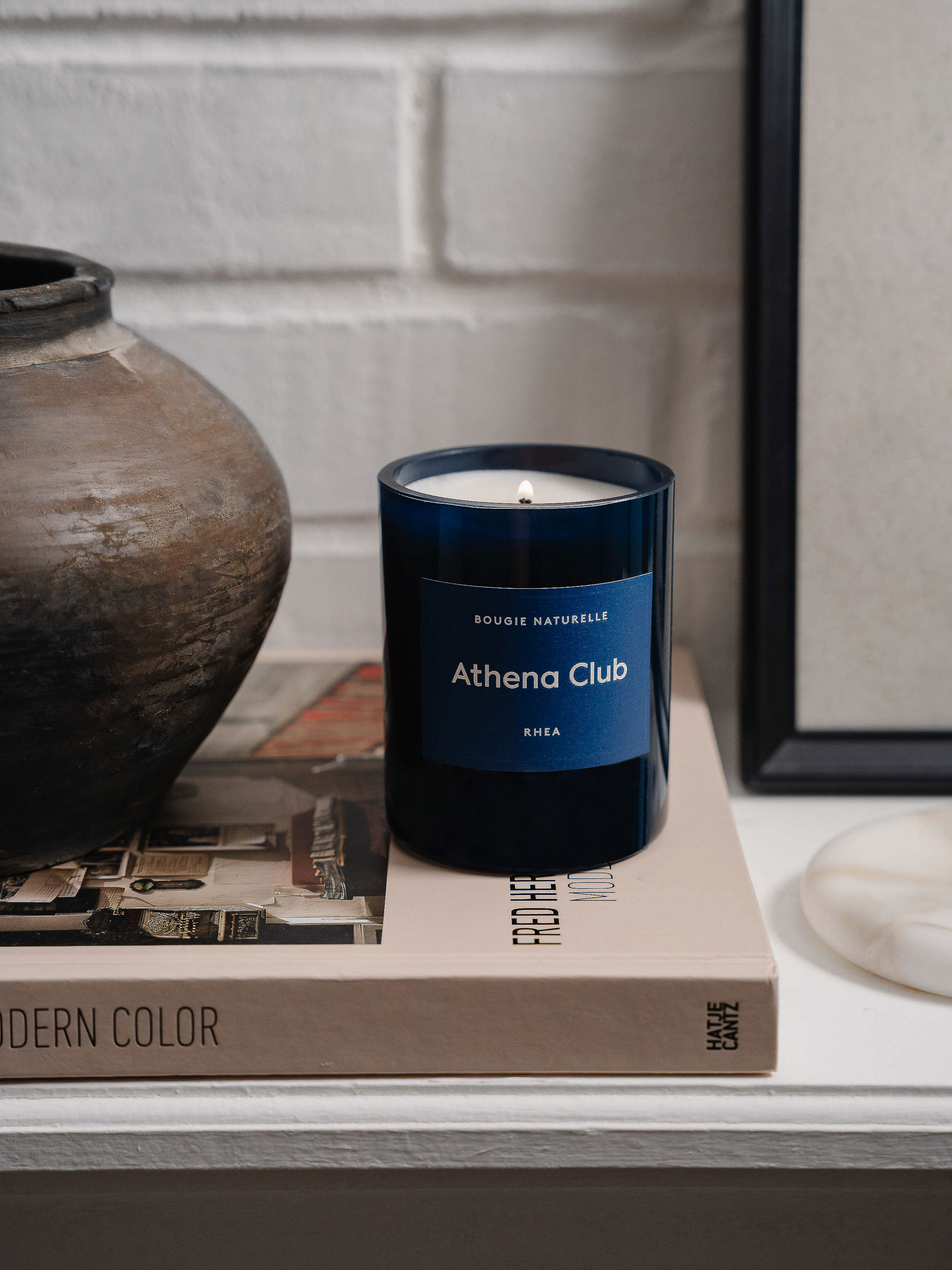 Dark blue Athena Club candle next to ceramic vase on books in front of a white brick wall and framed art.