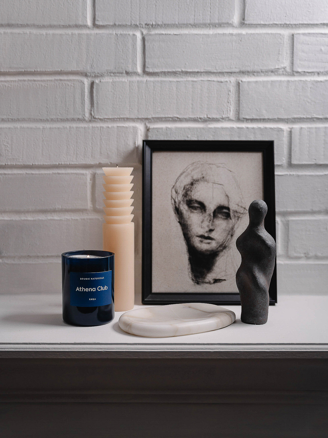Still life photography featuring a candle, framed portrait art, and a sculpture on a white mantel against a white brick wall.