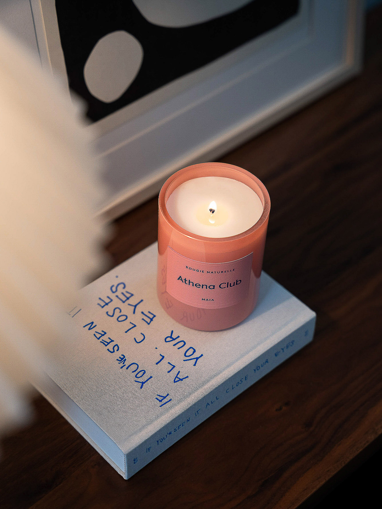 A lit pink candle from Athena Club sits atop a closed book with blue lettering on a dark wood surface, near framed art.