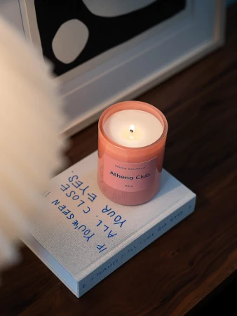 A lit pink candle from Athena Club sits atop a closed book with blue lettering on a dark wood surface, near framed art.