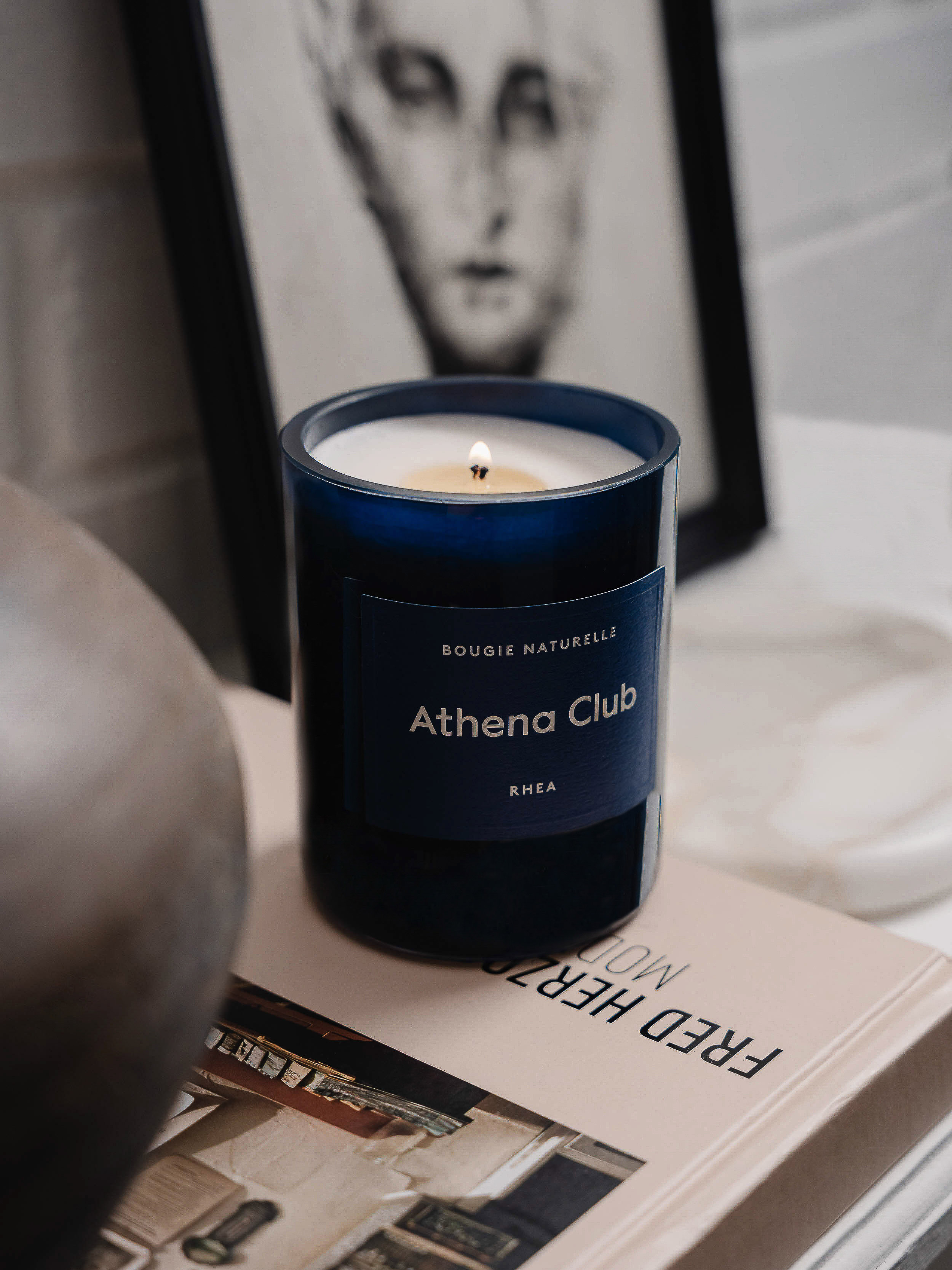 A lit blue candle with white wax and 'Athena Club' on the label sits on top of a book, with an art print in the background.