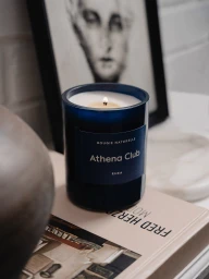 A lit Athena Club Rhea scented candle in a dark blue glass jar placed on a coffee table art book beside a framed art print.