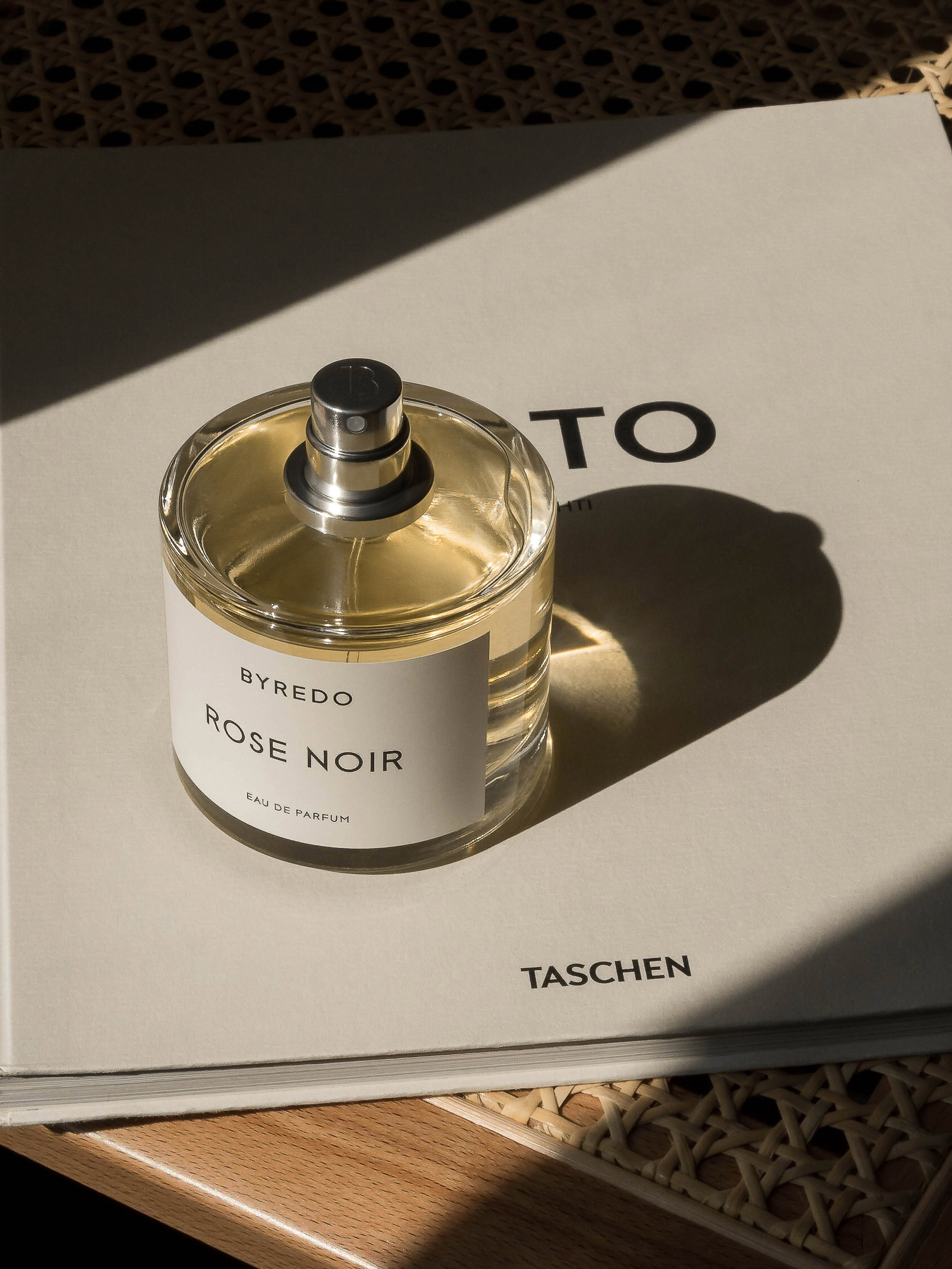 Byredo Rose Noir perfume bottle displayed on a hardcover book, casting soft shadows on a woven surface, evoking luxury and subtle sophistication.