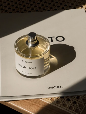 Byredo Rose Noir perfume bottle displayed on a hardcover book, casting soft shadows on a woven surface, evoking luxury and subtle sophistication.