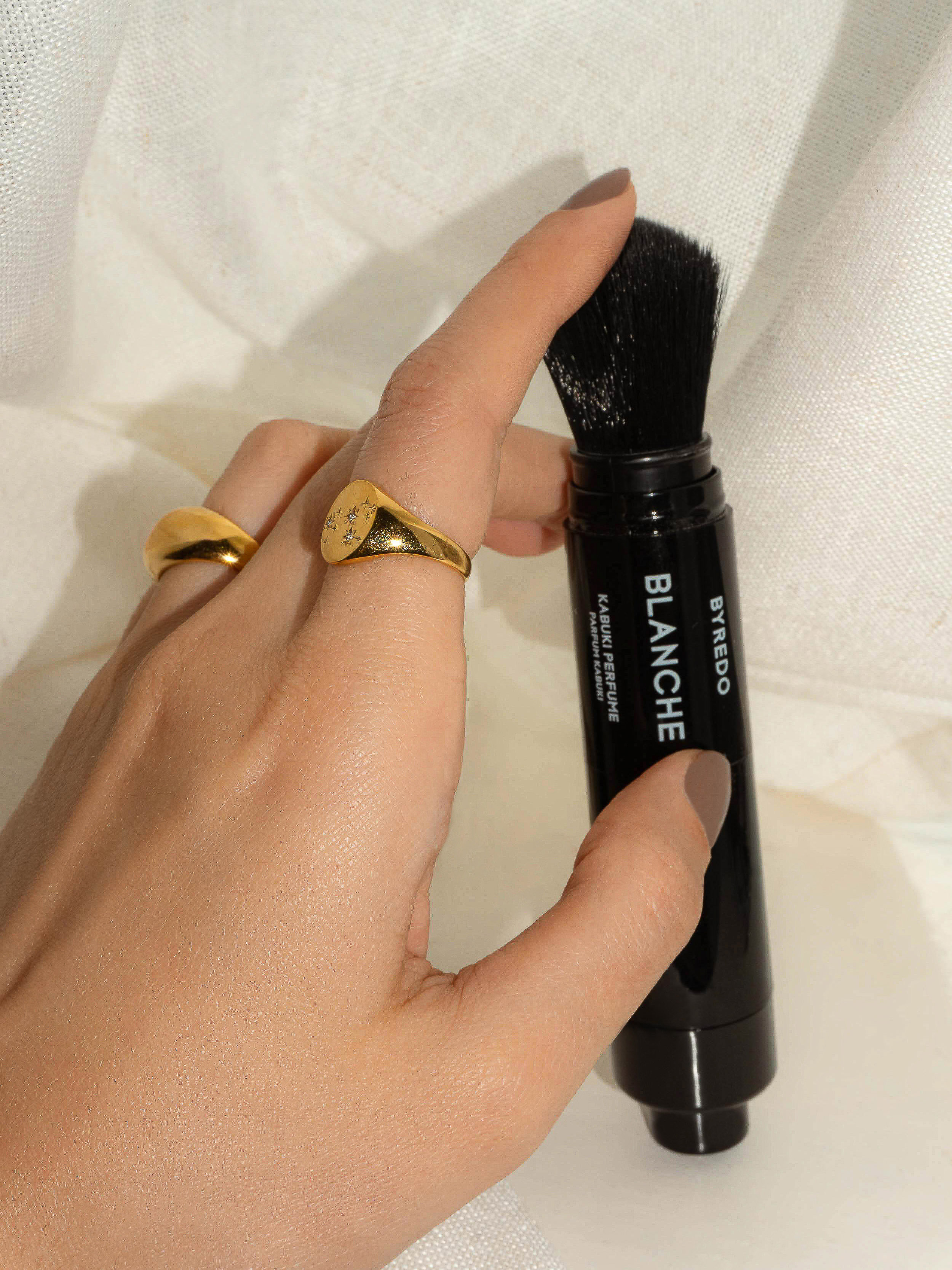 Hand holding Byredo Blanche Kabuki Perfume. Black container with brush applicator. Gold rings on fingers. Beauty and fashion product.
