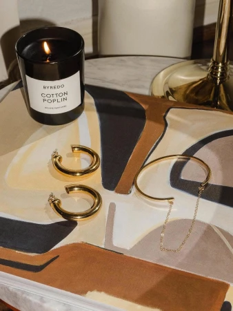 Elegant composition featuring gold jewelry, a Byredo candle, and an artful surface creates a luxurious and aesthetically pleasing scene.