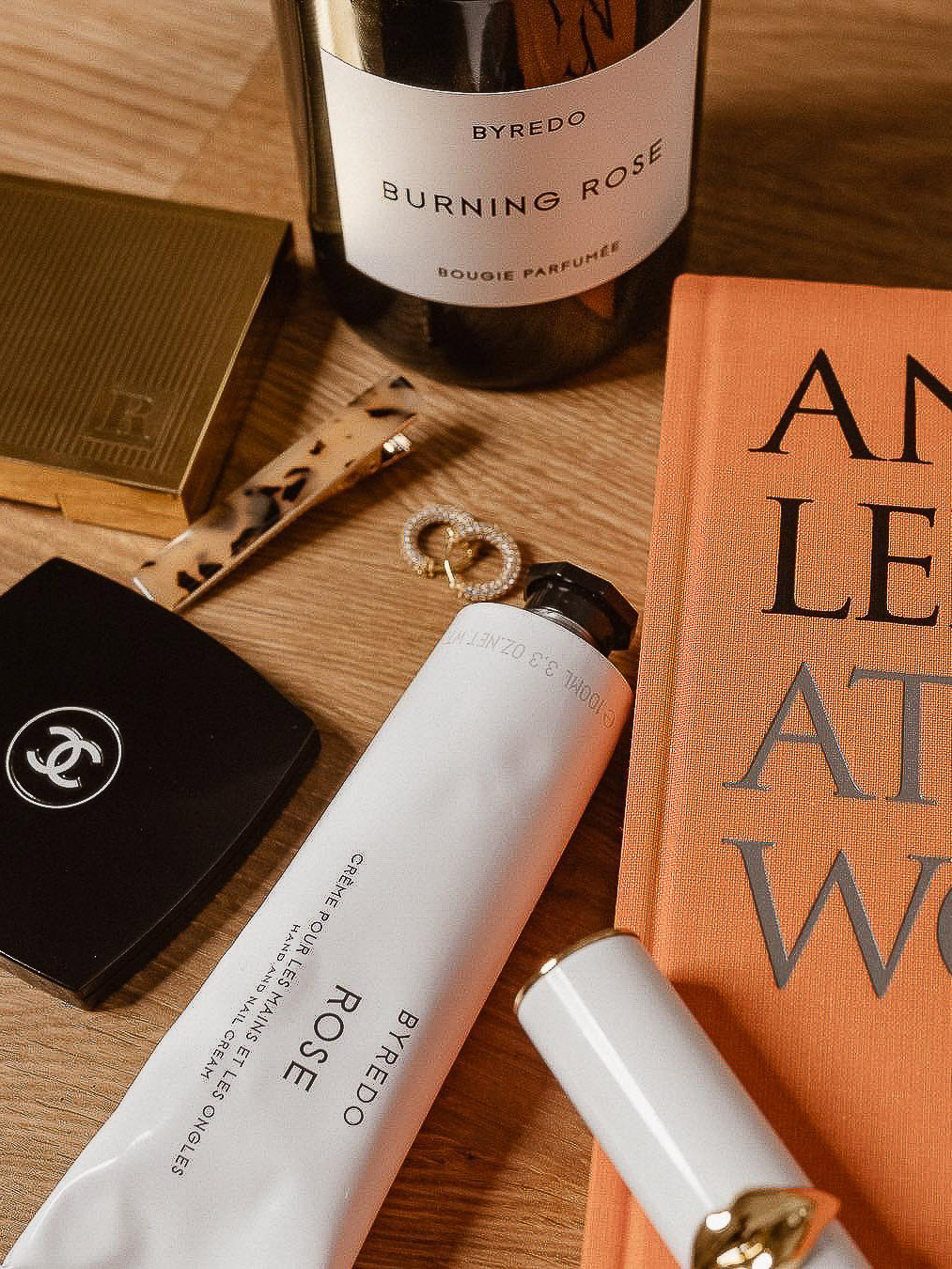 Still life shot of Byredo hand cream, candle, lipstick, Chanel compact, tortoiseshell hair clip, gold earrings, and a book.