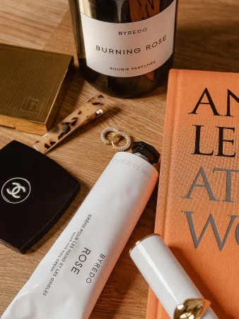 Still life shot of Byredo hand cream, candle, lipstick, Chanel compact, tortoiseshell hair clip, gold earrings, and a book.
