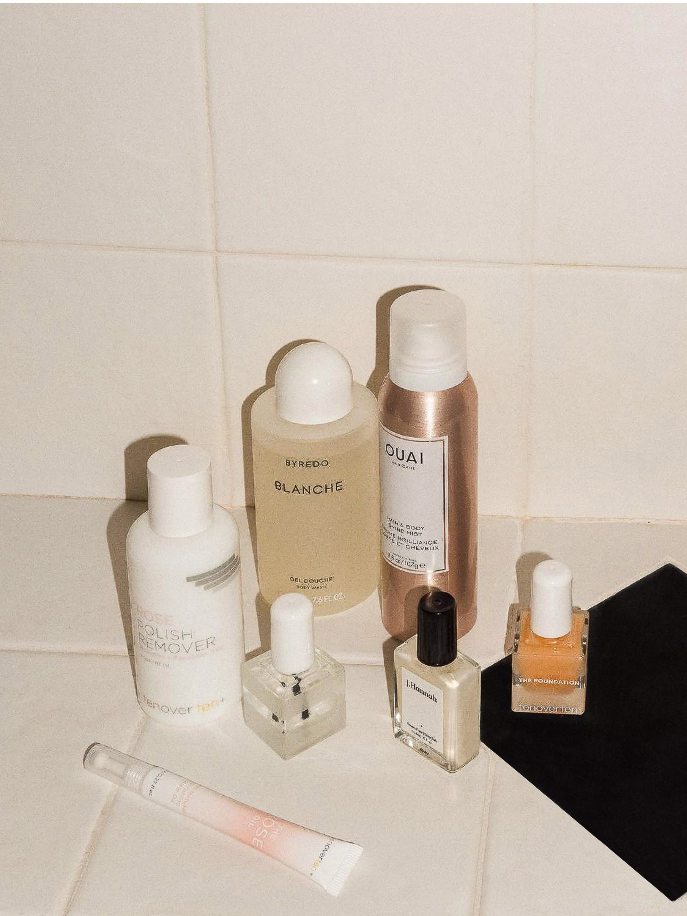 Cosmetic product arrangement on a white tiled surface, including nail polish, skin cream, and hair product bottles in a beauty product flatlay.