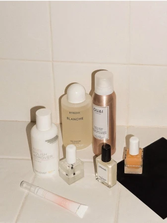 Cosmetic product arrangement on a white tiled surface, including nail polish, skin cream, and hair product bottles in a beauty product flatlay.
