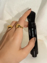 Hand wearing gold rings holding a black Byredo Blanche Kabuki Perfume brush against draped white fabric.