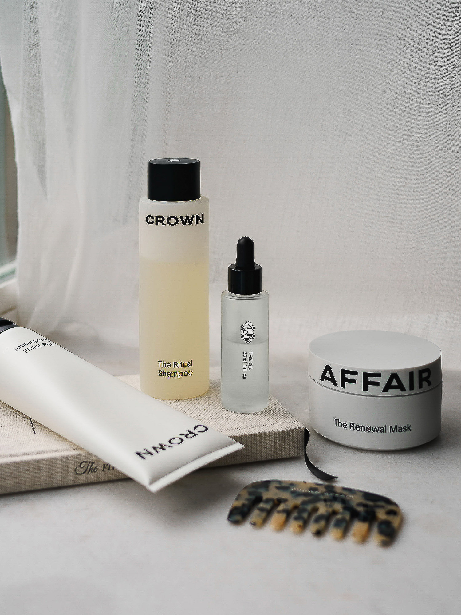 Still life of Crown and Affair beauty products including shampoo, renewal mask, and hair oil on a marble surface with comb.