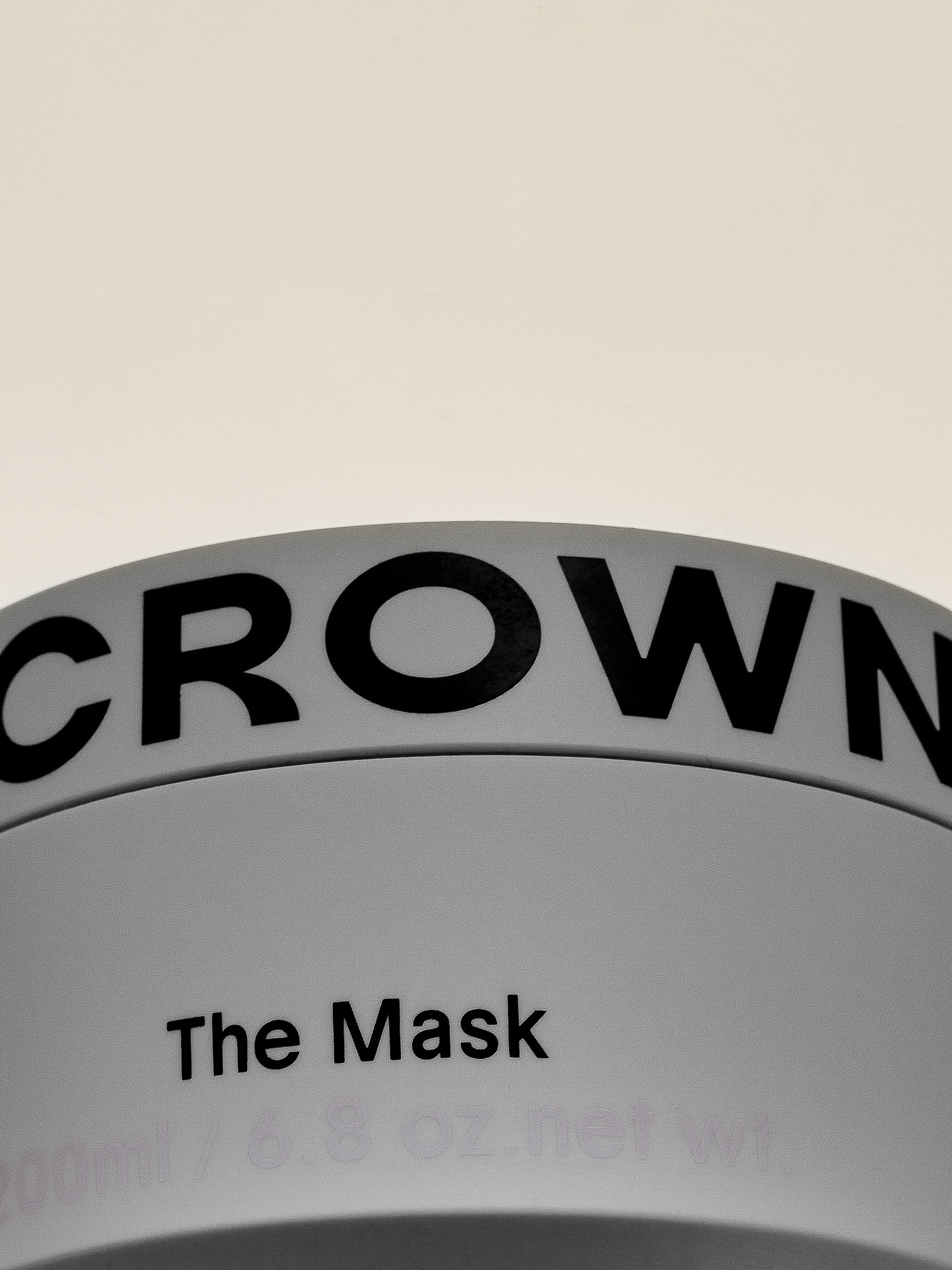 Close-up of a gray container labeled 'CROWN' and 'The Mask' in black lettering against a light beige backdrop, beauty product.