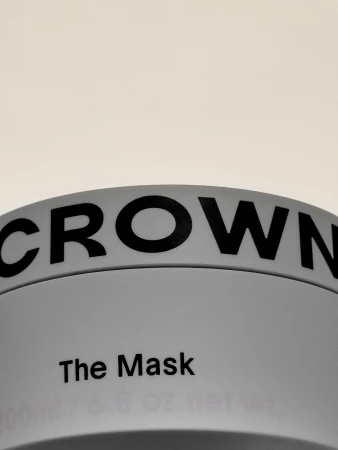 Close-up of a gray container labeled 'CROWN' and 'The Mask' in black lettering against a light beige backdrop, beauty product.