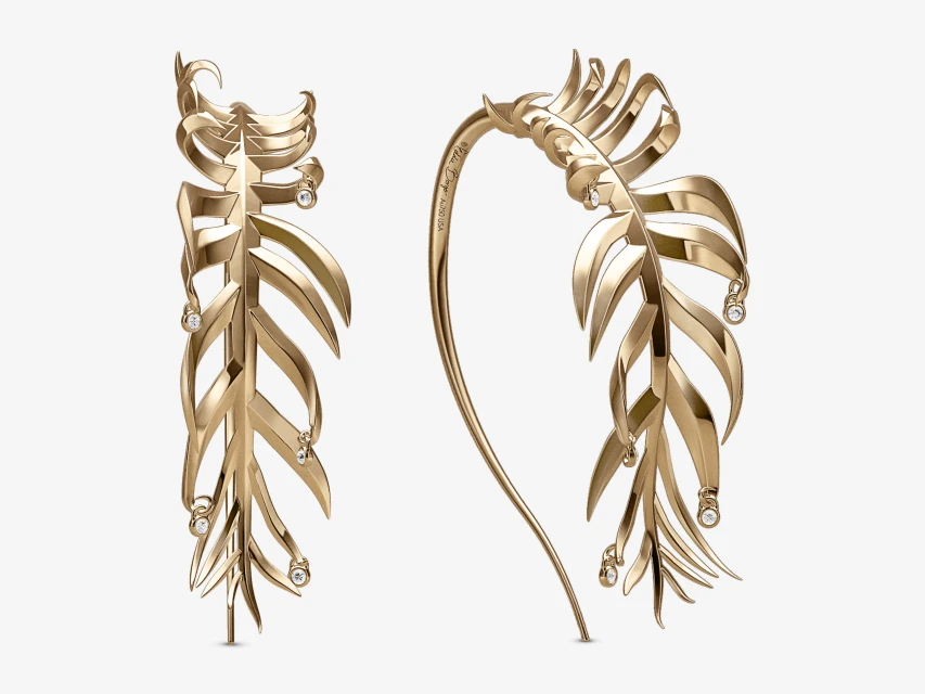 A pair of decorative gold hair accessories in the shape of a leaf, adorned with small diamonds, are displayed on a white backdrop.