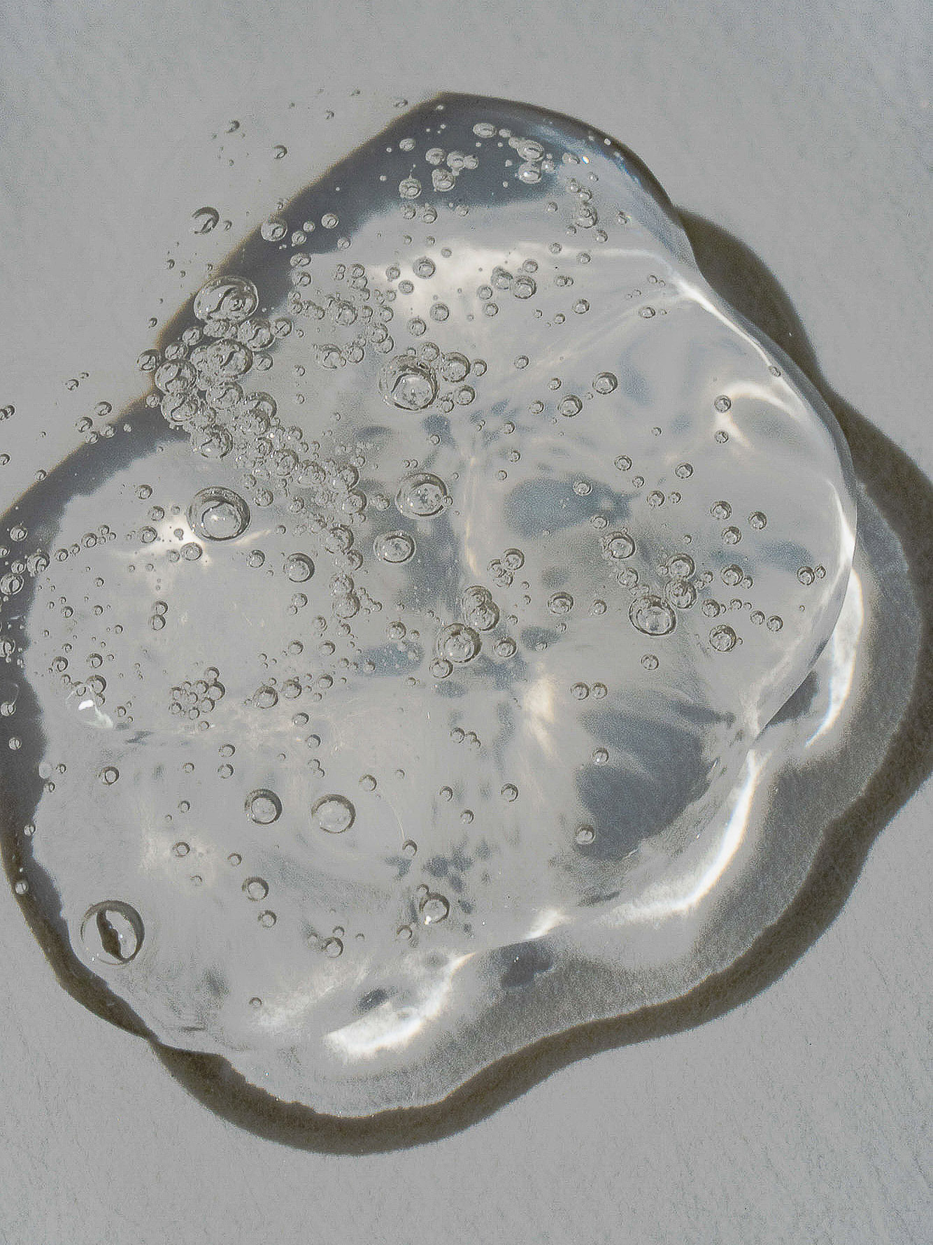 Clear cosmetic gel with bubbles on a gray surface. The gel has a thick, viscous texture and reflects light, indicating a hydrating formula.