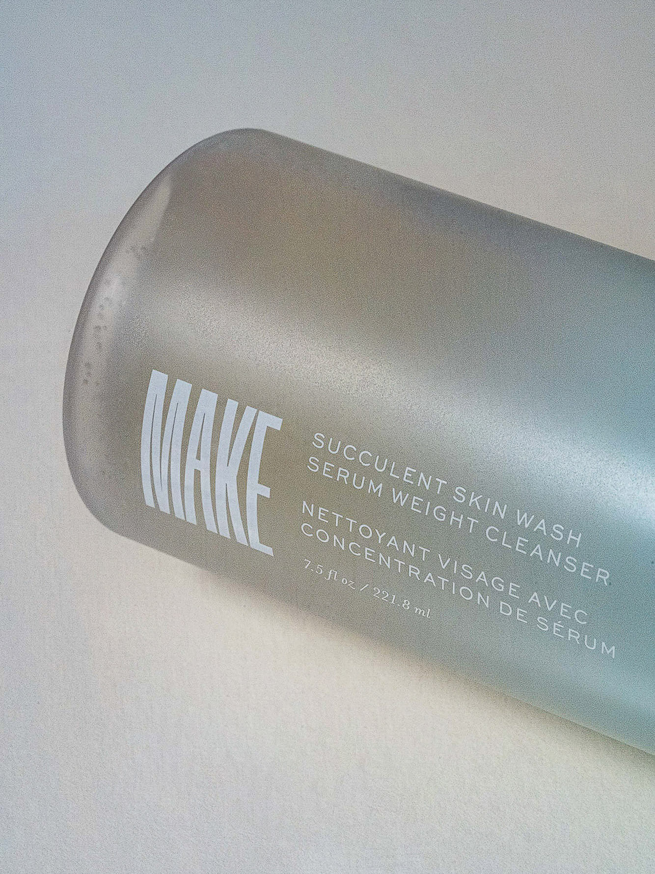 Close-up studio shot of a MAKE brand frosted bottle with text that reads 'Succulent Skin Wash Serum Weight Cleanser' on an off-white surface.