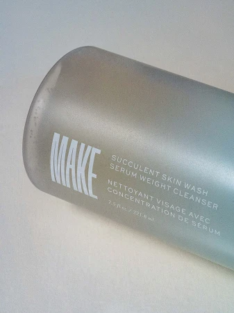 Close-up studio shot of a MAKE brand frosted bottle with text that reads 'Succulent Skin Wash Serum Weight Cleanser' on an off-white surface.