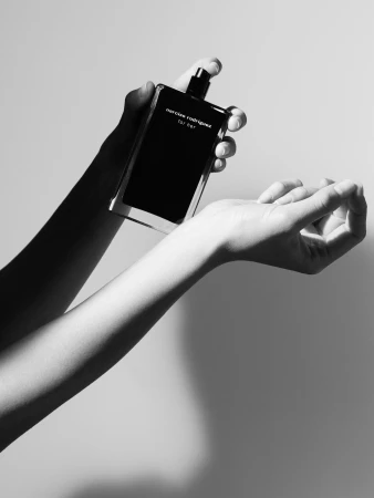 Black and white studio shot of woman's hands spraying Narciso Rodriguez For Her perfume onto her wrist; beauty product.