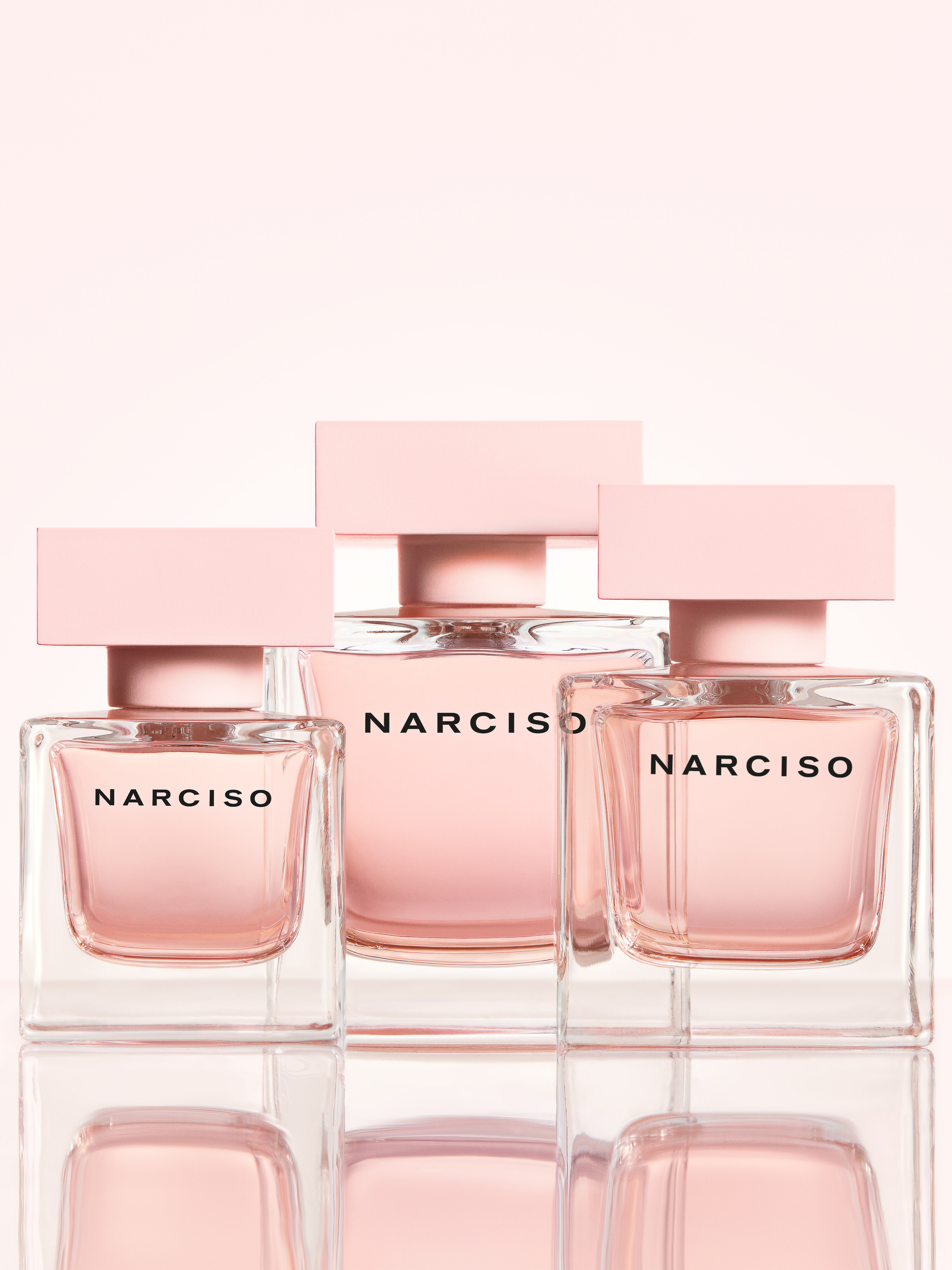 Three square-shaped Narciso Rodriguez perfume bottles in varying sizes sit on a glossy surface against a soft pink gradient background.