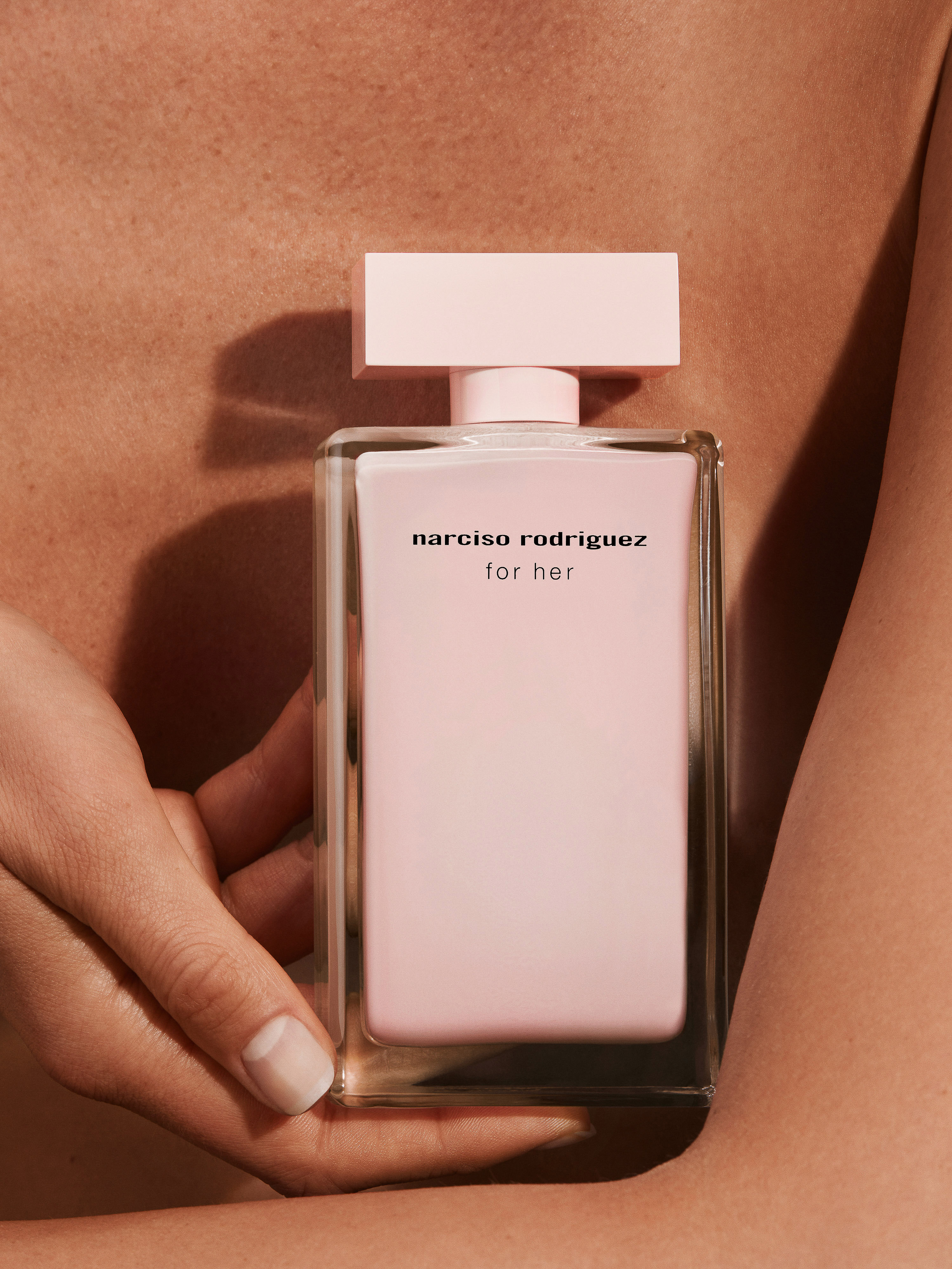 Close-up studio shot of a Narciso Rodriguez For Her perfume bottle held by a person, showcasing its minimalist design and soft pink hue.