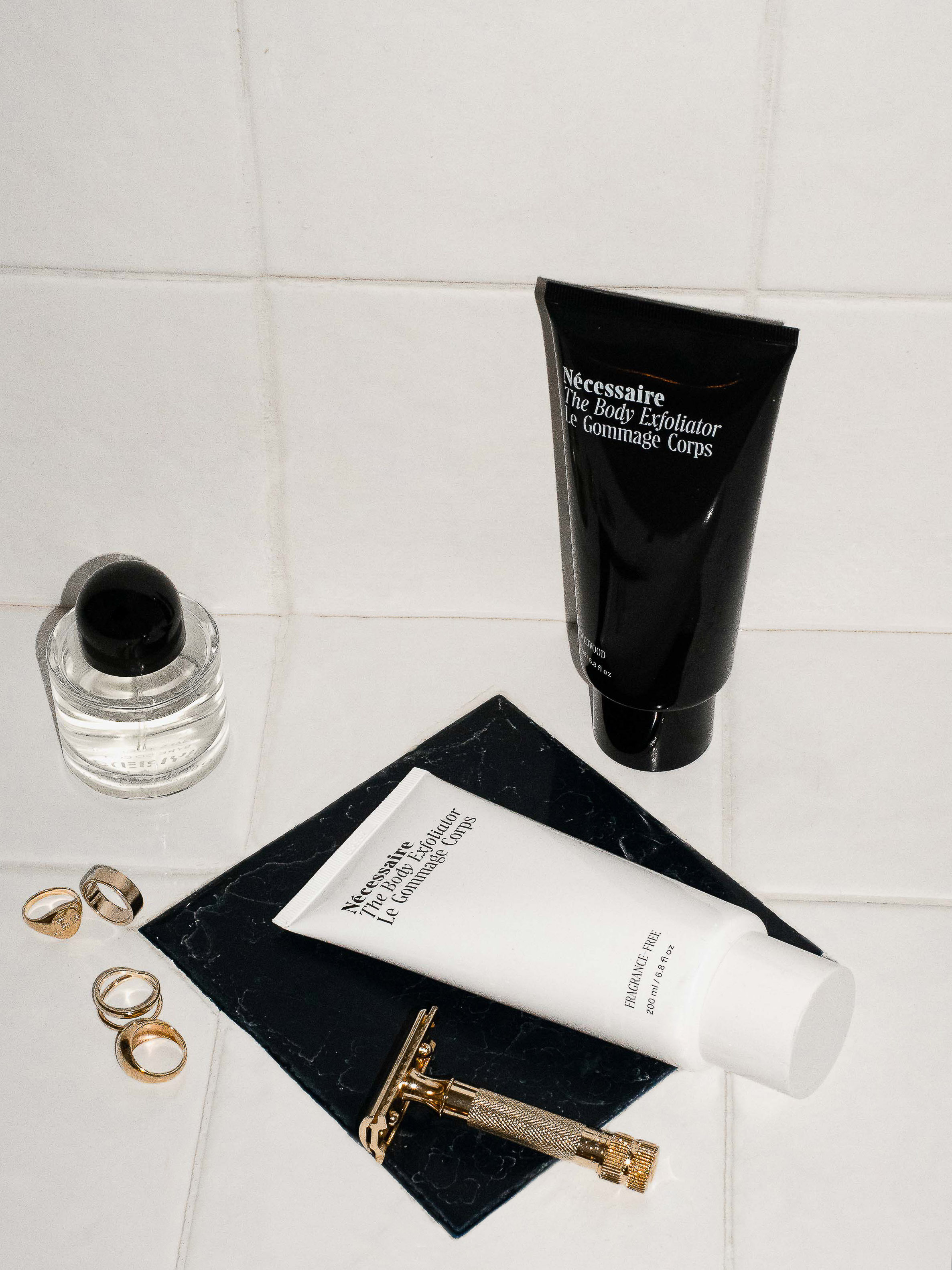 Cosmetic product arrangement featuring a black tube, white bottle, clear bottle, and safety razor on a black tile surface.