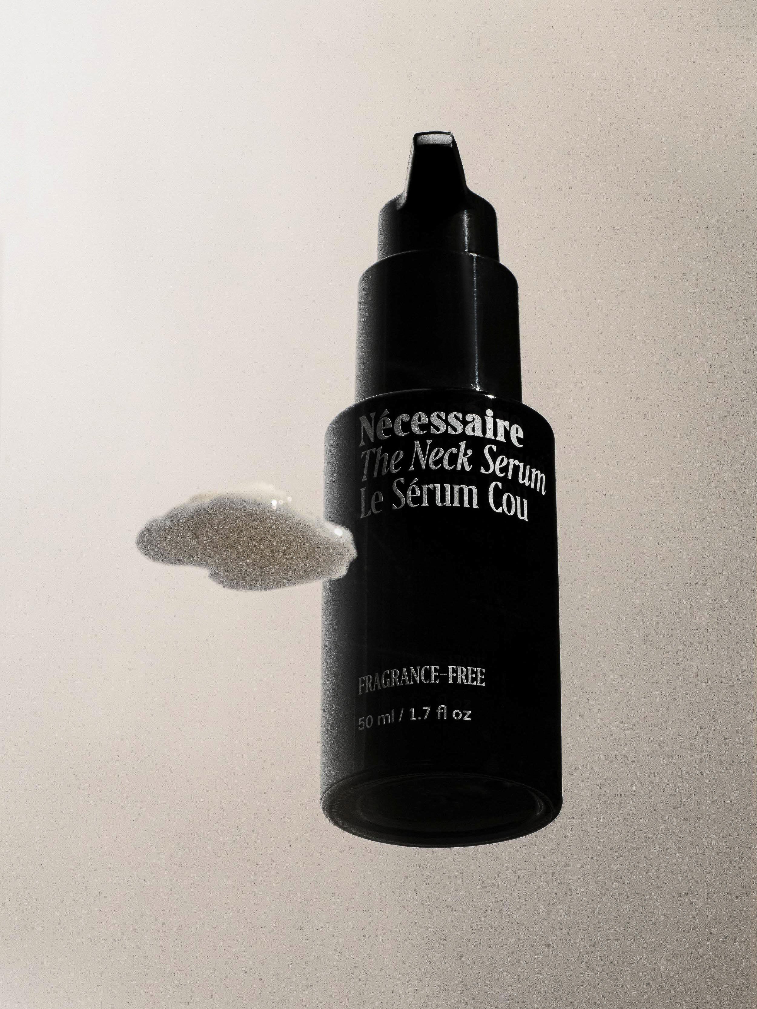 A bottle of Necessaire The Neck Serum sits next to a dollop of the white cream on a white background, in a product shot.