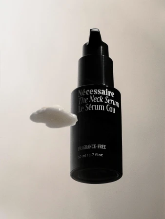 A bottle of Necessaire The Neck Serum sits next to a dollop of the white cream on a white background, in a product shot.