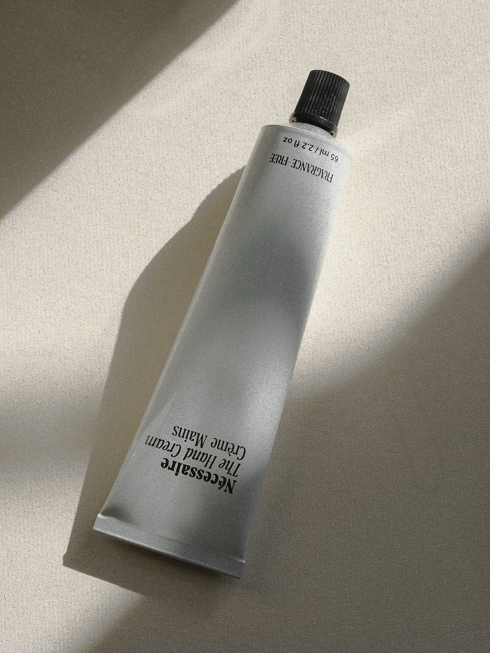 Minimalist still life of a gray tube of Nécessaire The Hand Cream on a cream-colored surface with a soft, diffused light and shadow.