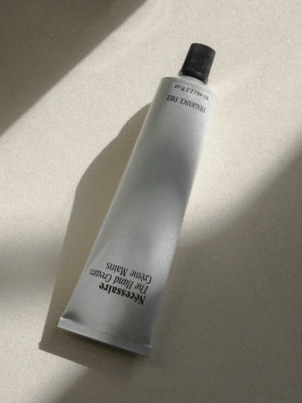 Minimalist still life of a gray tube of Nécessaire The Hand Cream on a cream-colored surface with a soft, diffused light and shadow.