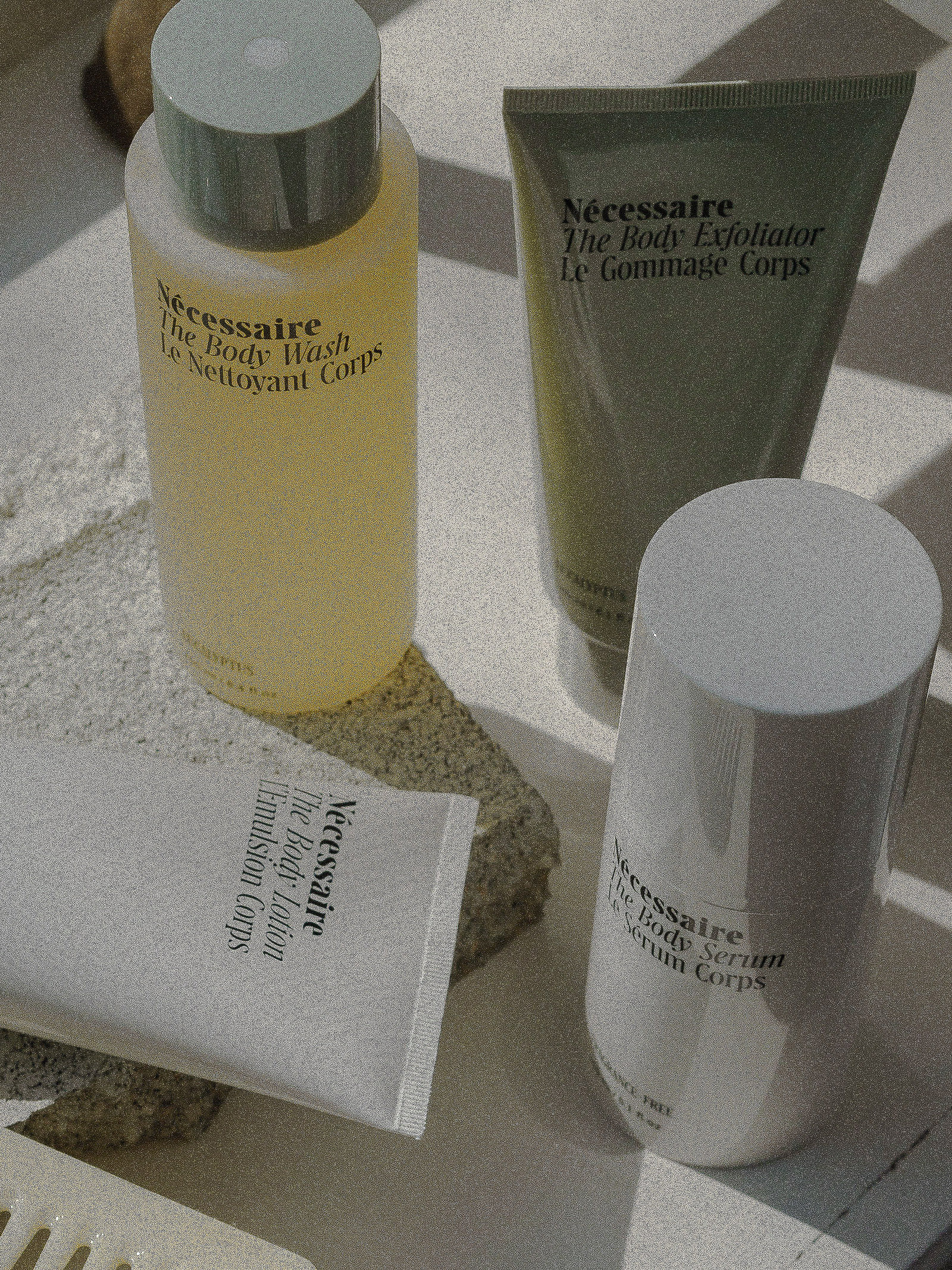 Close-up of Nécessaire body care products including body wash, exfoliator, lotion, and serum, arranged on a textured surface in soft lighting.
