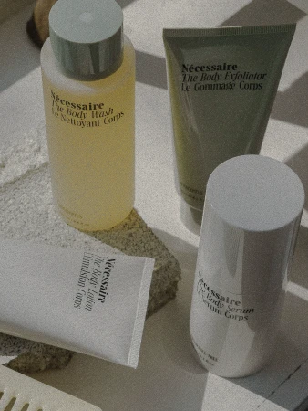 Close-up of Nécessaire body care products including body wash, exfoliator, lotion, and serum, arranged on a textured surface in soft lighting.