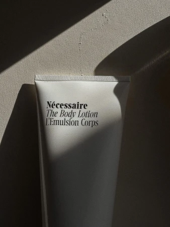 Close-up of a white tube of Nécessaire The Body Lotion L'Emulsion Corps against a neutral background with strong shadows.
