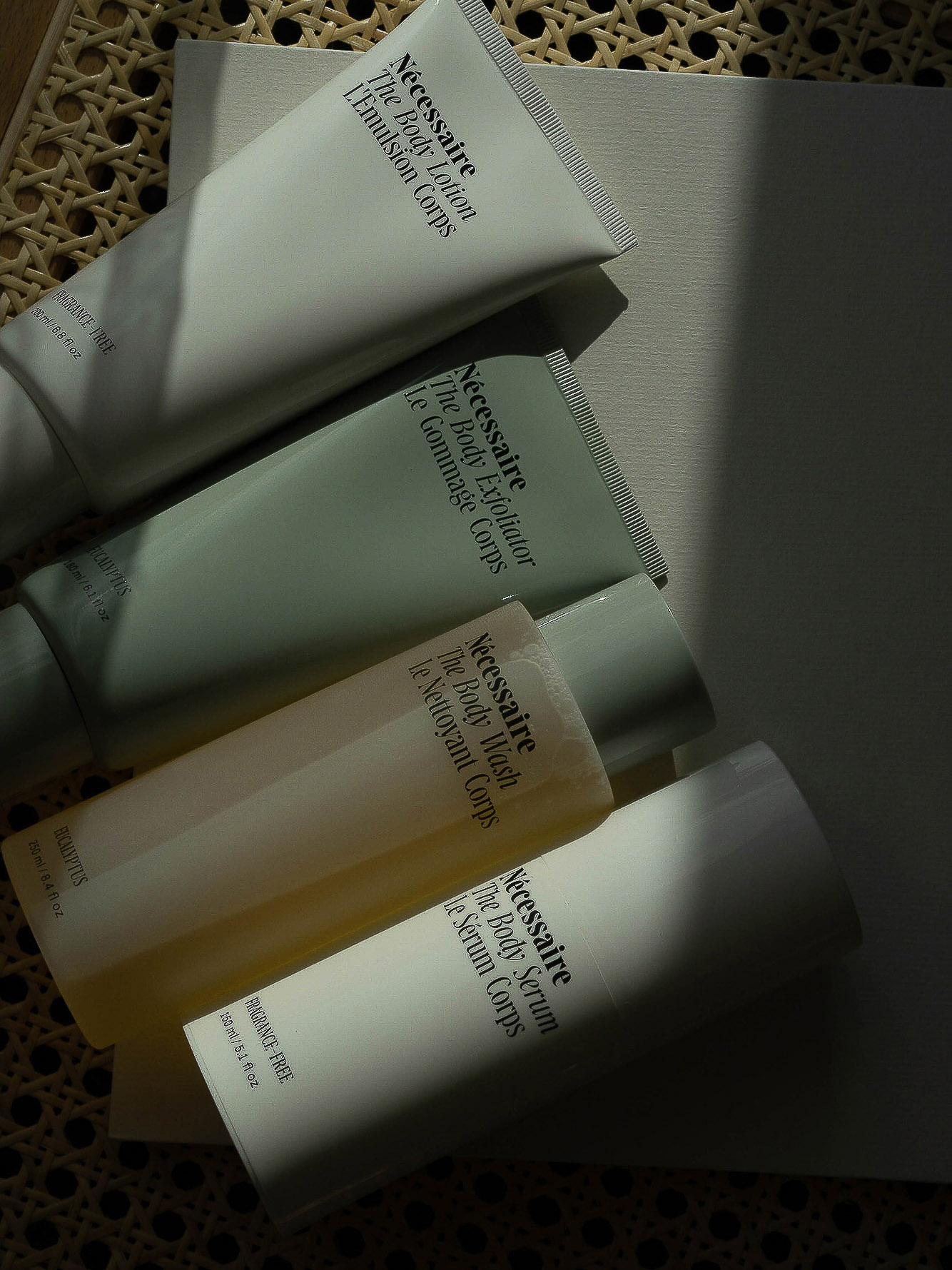 Four tubes of Nécessaire body lotion, exfoliator, wash, and serum are arranged on a white surface against a woven background.