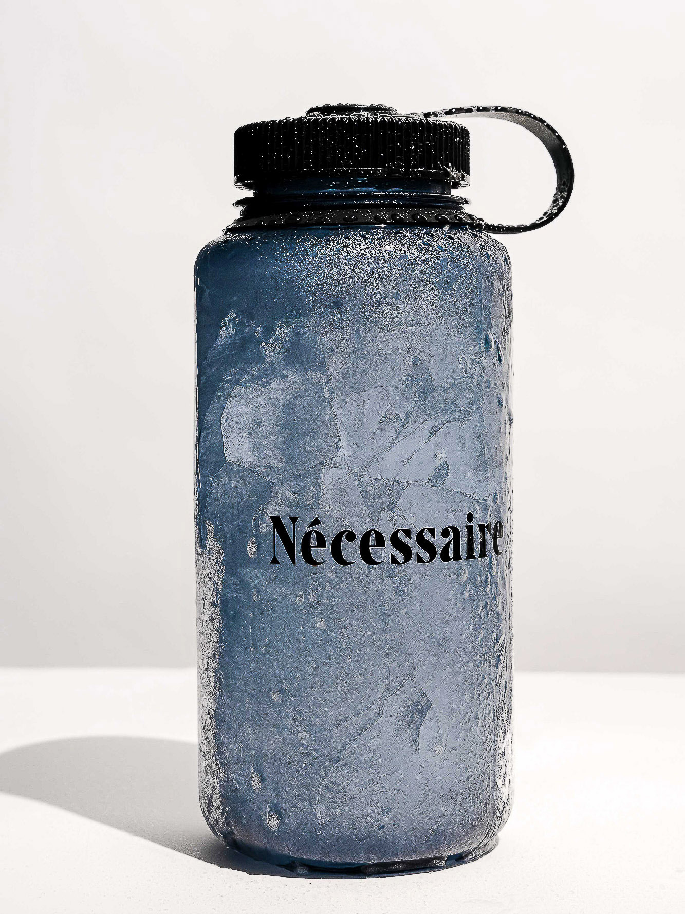 Blue water bottle filled with ice and condensation on a white surface, featuring the word "Nécessaire" in black text.
