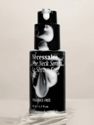 Black bottle of Nécessaire The Neck Serum covered in smeared white cream on a pale seamless surface.