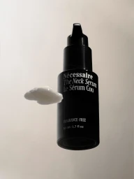 Black bottle of Nécessaire The Neck Serum beside a swatch of white cosmetic cream against a light grey background.