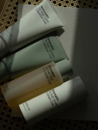 Overhead view of four Necessaire body care bottles and tubes arranged diagonally across paper and woven cane with dramatic shadows.