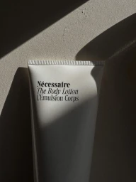 White tube of Nécessaire The Body Lotion highlighted with strong diagonal shadows against a neutral textured surface.